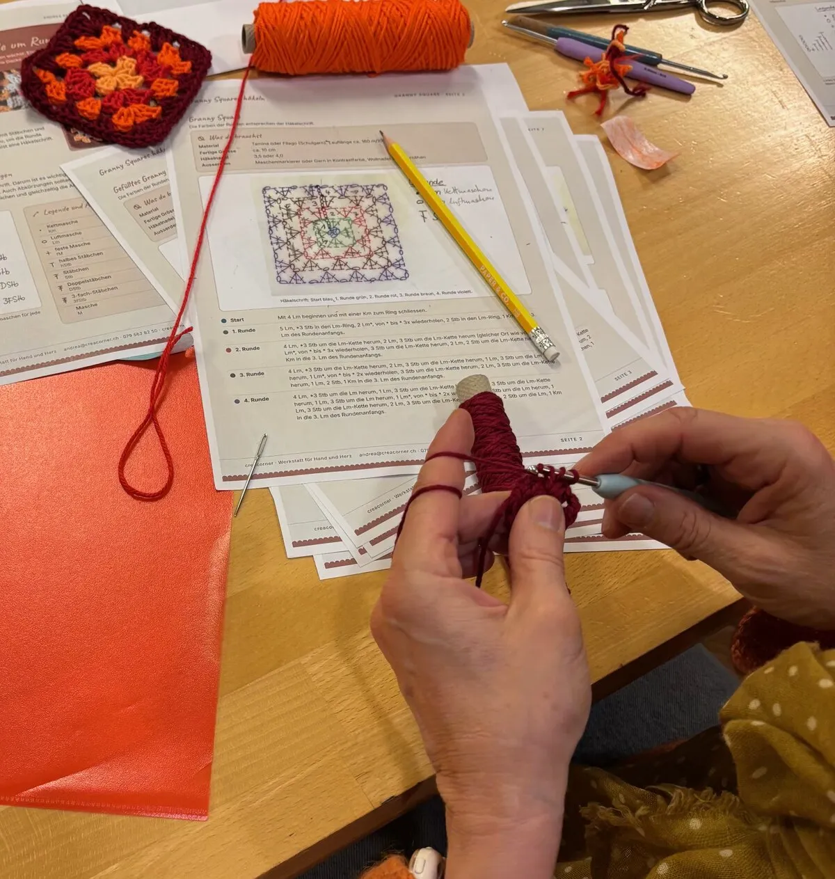 Zwei Hände häkeln mit dunkelrotem Garn über der Anleitung für Granny Squares
